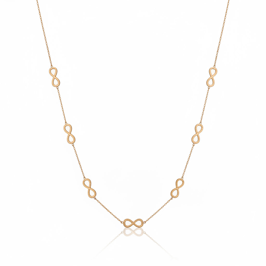 A delicate gold chain necklace featuring multiple small infinity loop charms spaced along the chain, displayed on a reflective surface with soft window shadow projections.