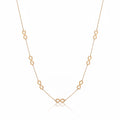 A delicate gold chain necklace featuring multiple small infinity loop charms spaced along the chain, displayed on a reflective surface with soft window shadow projections.